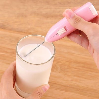 main utilisant un mousseur a lait rose pour mousser du lait dans un verre, avec un arrière-plan en bois naturel