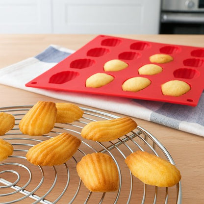 des madeleines fraîchement cuites disposées sur une grille de refroidissement avec un moule en silicone rouge contenant des madeleines encore à cuir