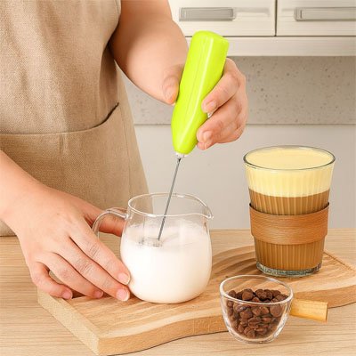 main utilisant Mousseur a lait vert pour faire mousser du lait dans un pichet avec une tasse de café en arrière-plan