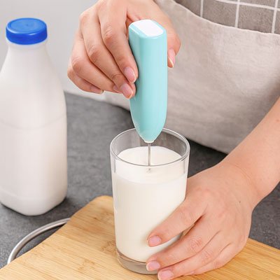 main utilisant un Mousseur a lait bleu pour faire mousser du lait dans un verre, avec une bouteille de lait à côté
