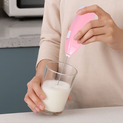 main utilisant un Mousseur a lait rose pour faire mousser du lait dans un verre, avec une cuisine moderne en arrière-plan