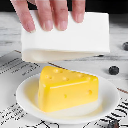 Une main soulève un moule à gâteau blanc pour révéler un gâteau en forme de part de fromage jaune posé sur une petite assiette